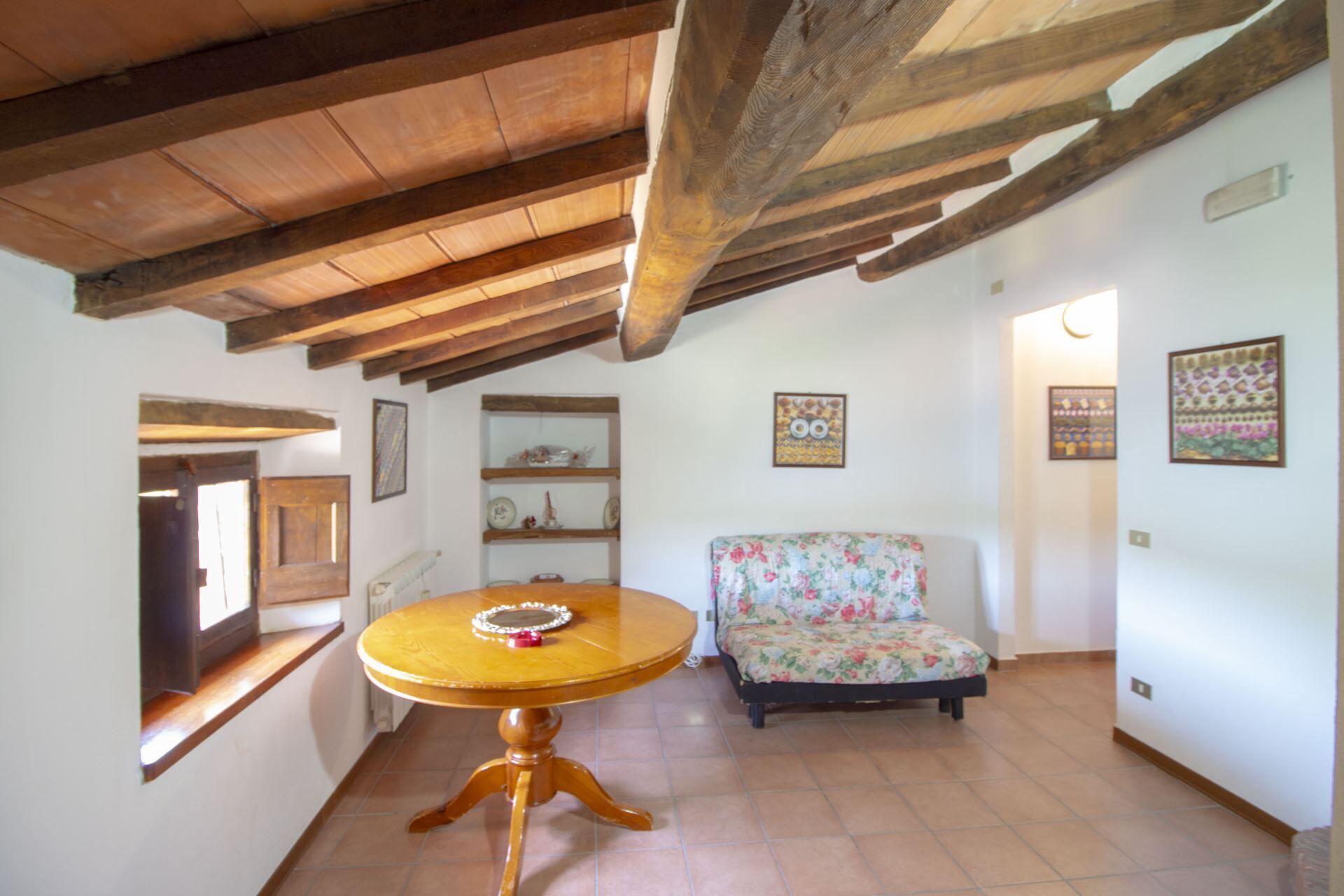 A room with a wooden ceiling, a round table with a decorative item, a floral sofa, and shelves with various items.