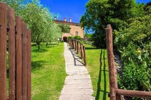 Ein Steinweg führt zu einem Steinhaus mit Backsteinfassade und Schornsteinen, umgeben von Grünflächen und einem Holzzaun.