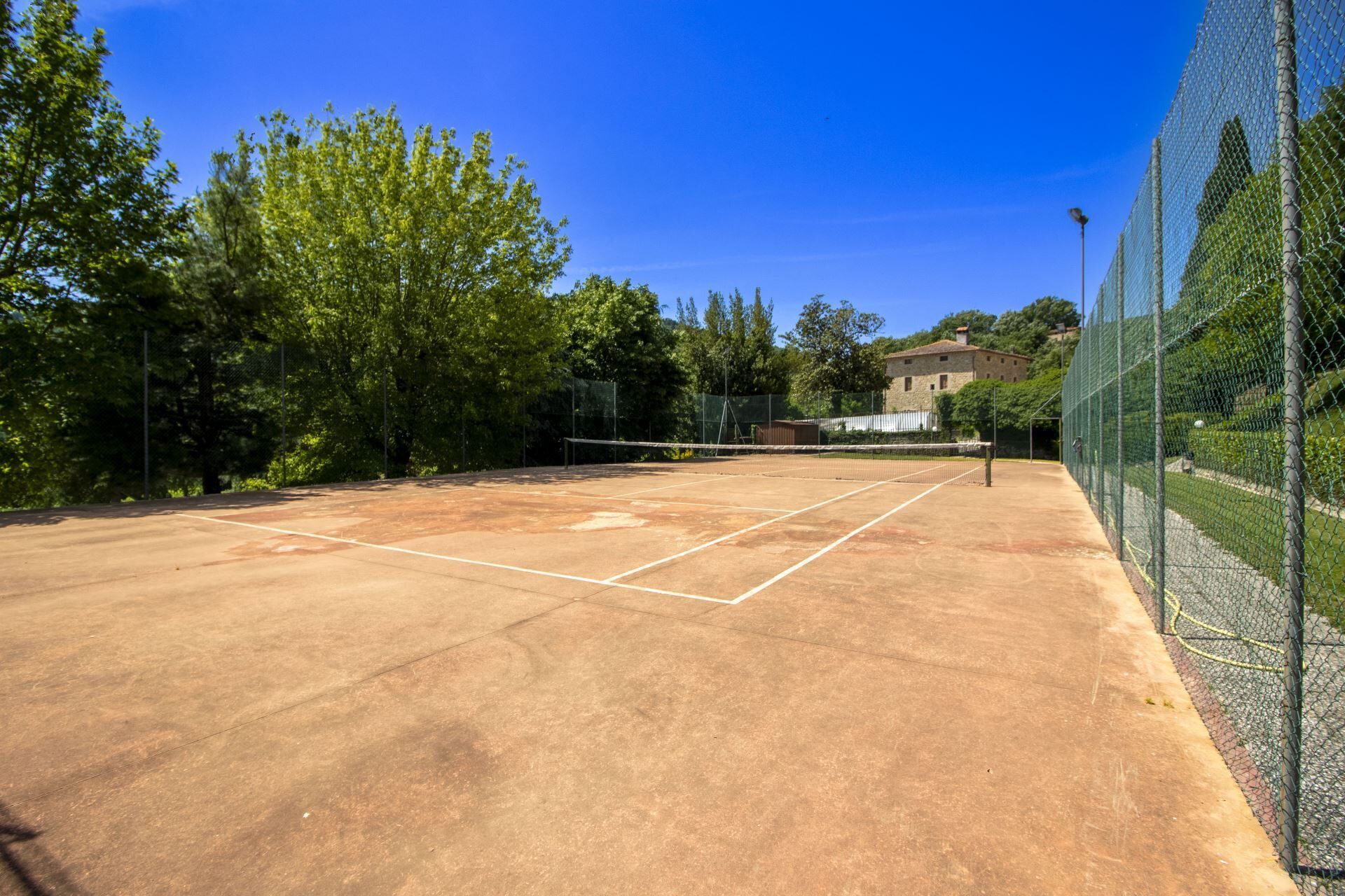 An outdoor tennis court with a net, surrounded by trees and a building in the background.