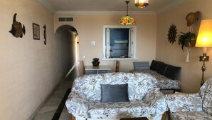 Un salon chaleureux avec un canapé à motifs floraux, un fauteuil assorti, une petite table, une valise et une fenêtre donnant sur la cuisine.