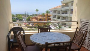Un balcon avec une table ronde et quatre chaises en bois, donnant sur un quartier résidentiel avec des immeubles et un ciel dégagé.