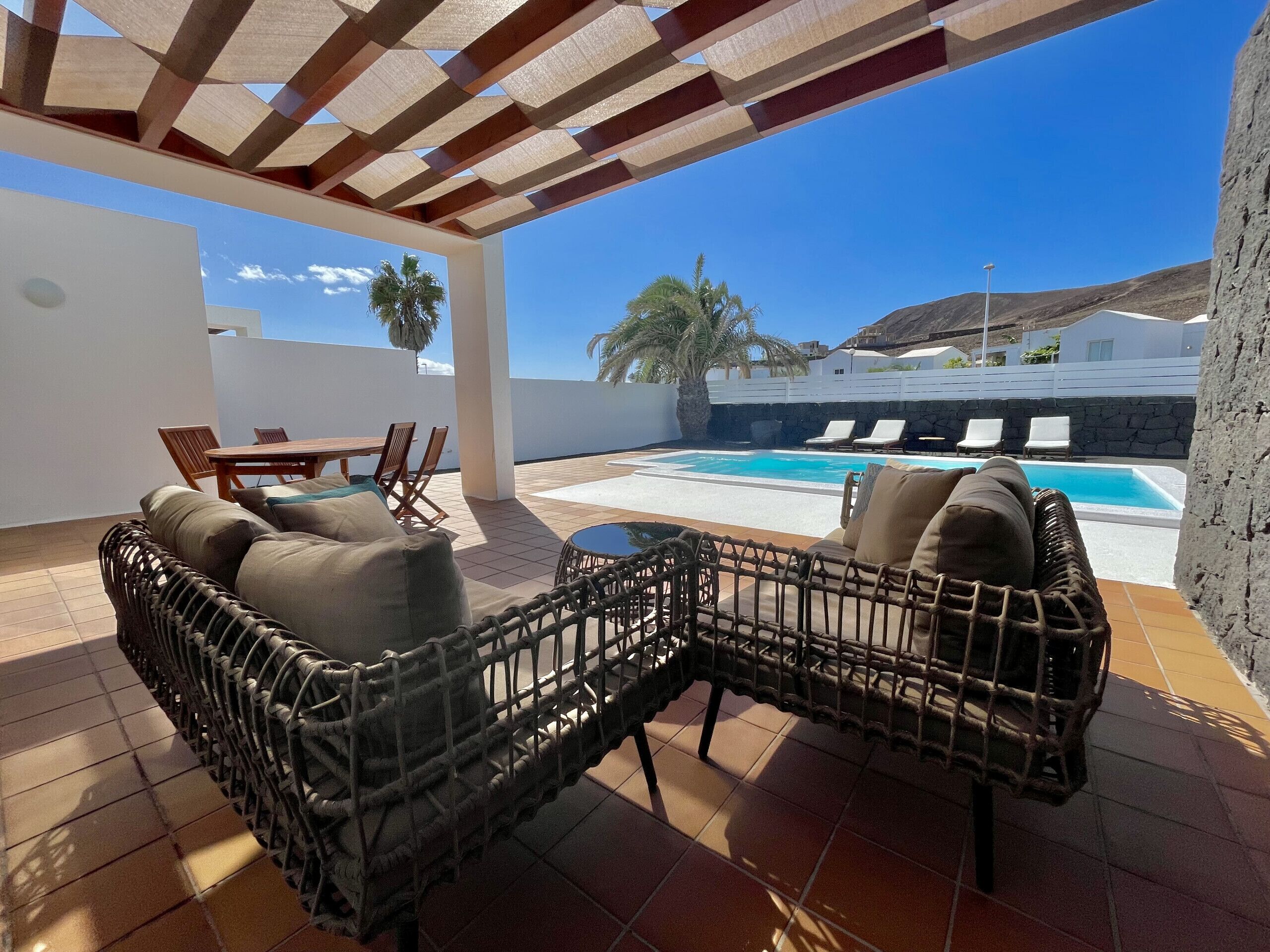 A covered outdoor seating area with wicker furniture, a pool, and a view of a white building and palm trees.