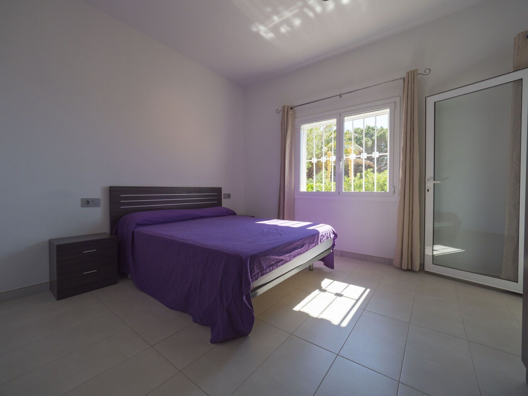 A bedroom with a bed, a nightstand, a sliding glass door, and a view of greenery outside.