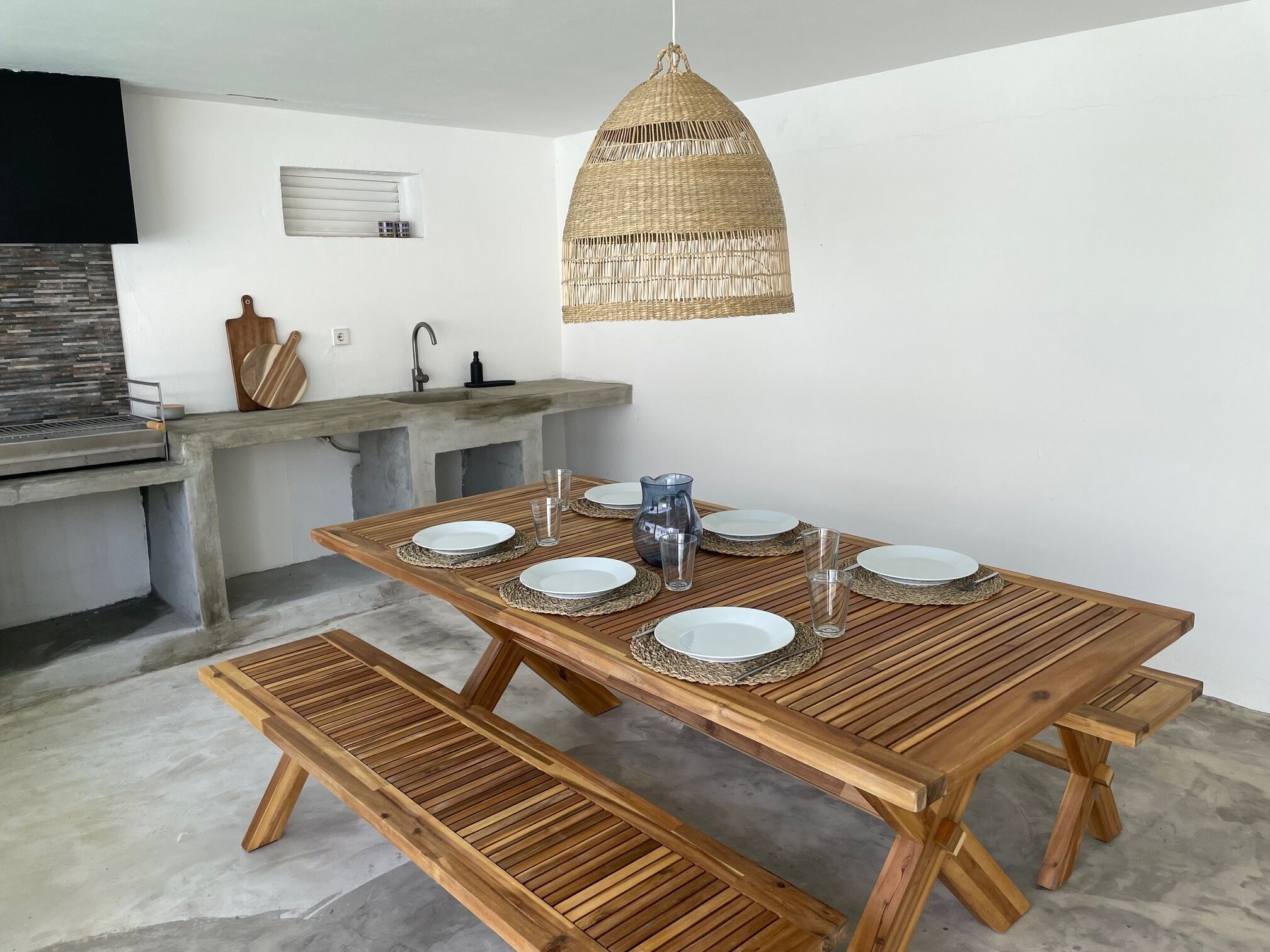 A wooden dining table with benches, a hanging rattan light fixture, and a concrete counter with a sink.