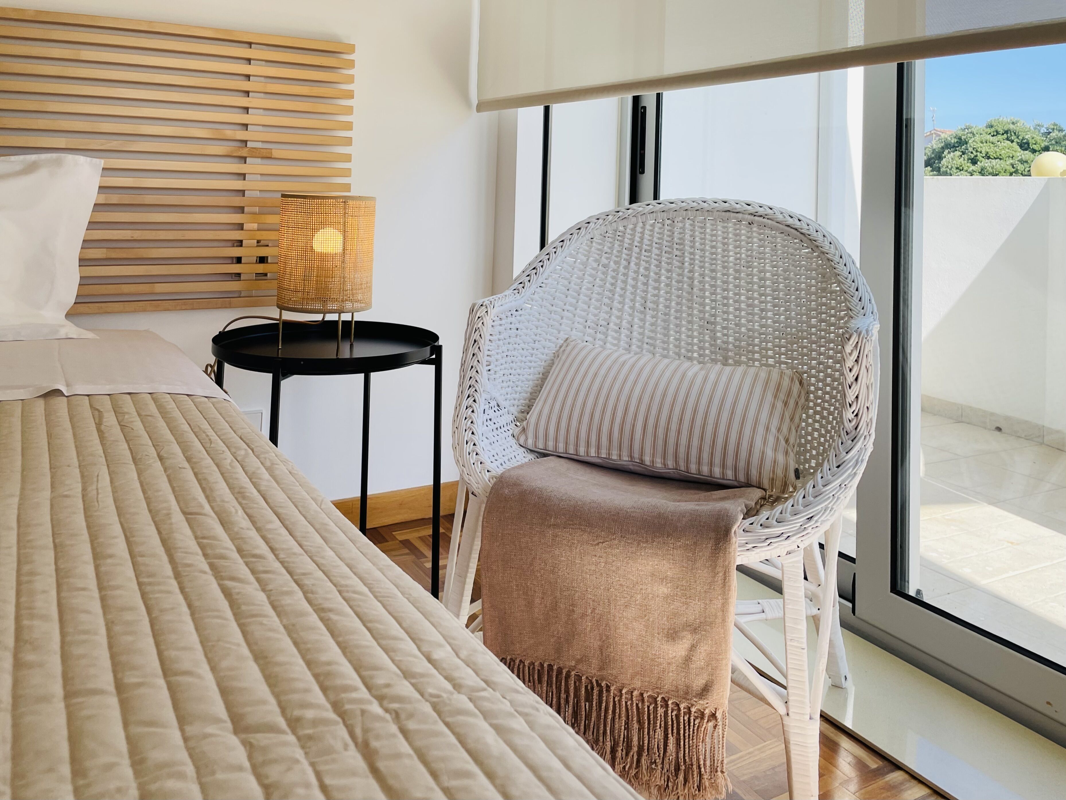 A modern bedroom with a large bed, a wicker chair with a cushion, a small table with a lamp, and a view of the outdoors through a large window.