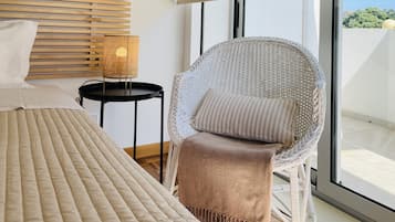 A modern bedroom with a large bed, a wicker chair with a cushion, a small table with a lamp, and a view of the outdoors through a large window.