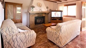 A living room with two patterned sofas, a fireplace, a television, and wooden beams on the ceiling. - Calas - Javea, Costa Blanca (Javea)