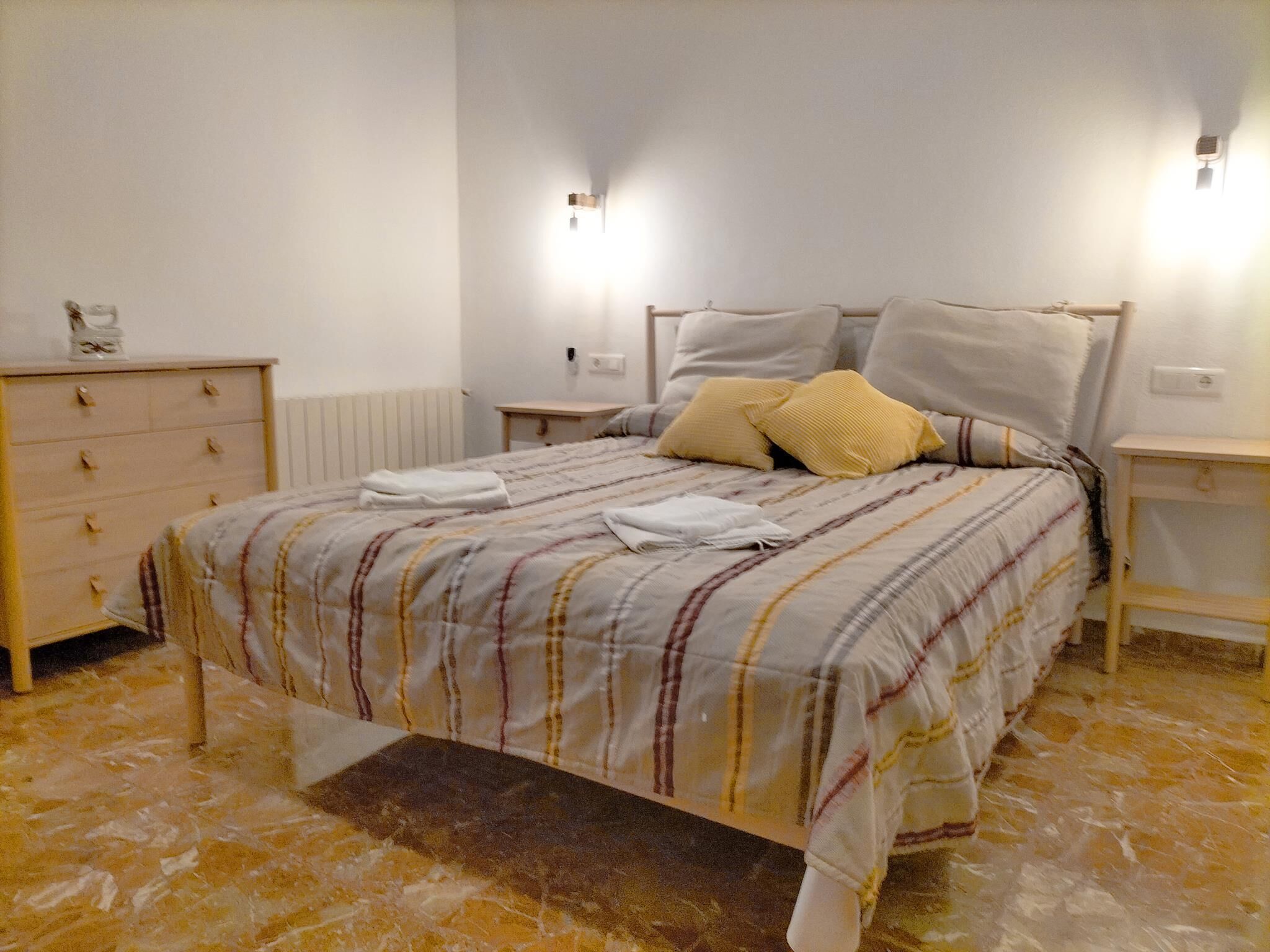 A bedroom with a bed, a dresser, two bedside tables, and a wall-mounted light fixture.