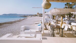 A beachside restaurant with white outdoor seating, a large hanging light fixture, and a view of the sea and distant buildings. - Calas - Javea, Costa Blanca (Javea)