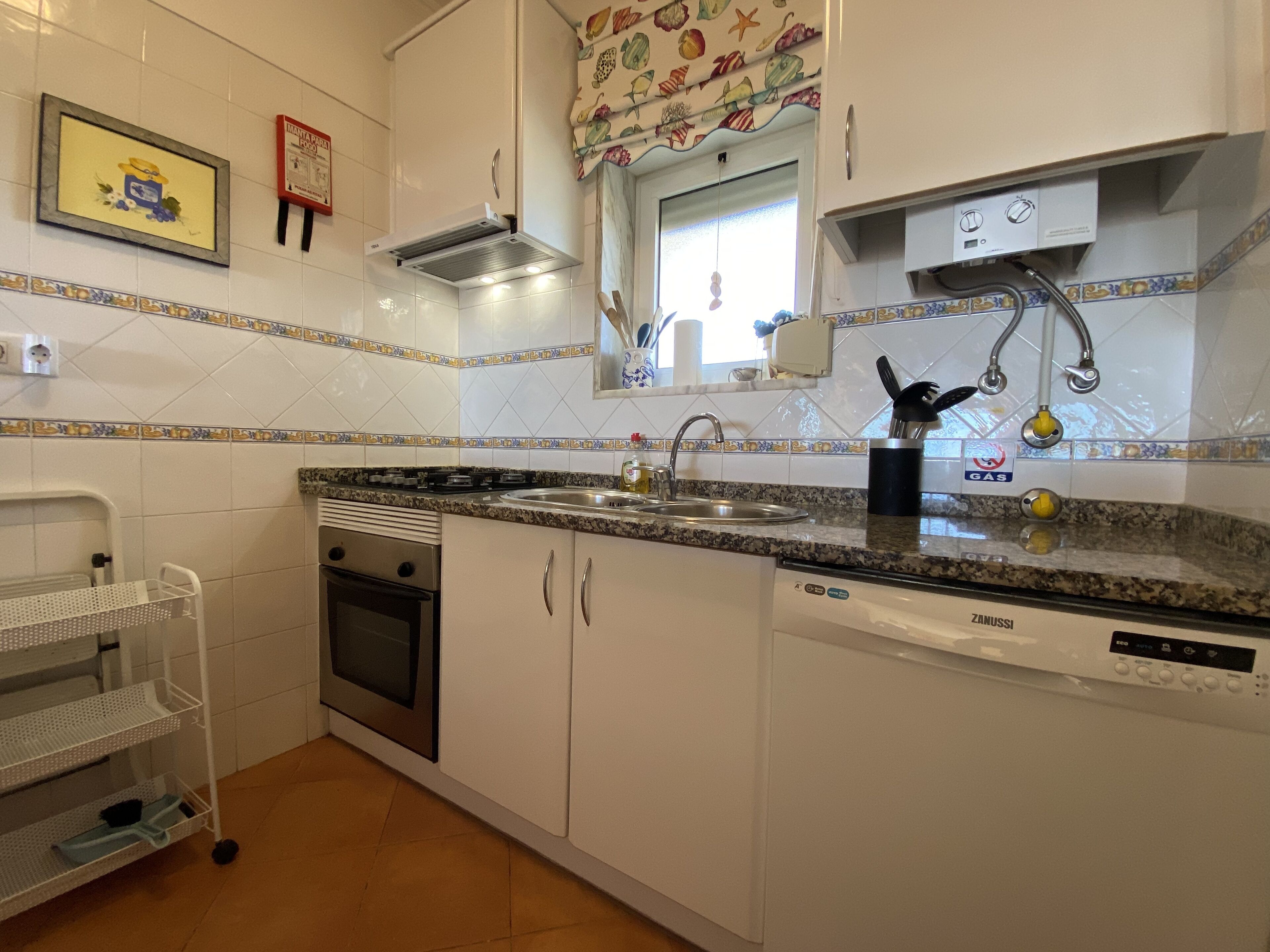 A kitchen with white cabinets, a granite countertop, a stove, an oven, a sink, and a dishwasher. There is a window above the sink, a gas stove, and a metal rack with dishes.