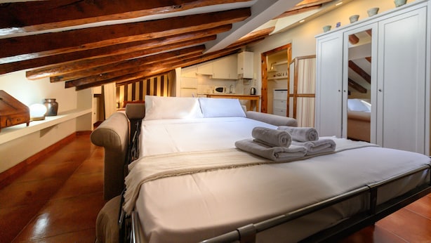 Un dormitorio con cama, techo de madera, armario blanco y una cocina al fondo.