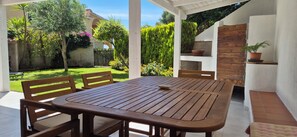 Un patio al aire libre con una mesa y sillas de madera, rodeado por un jardín con diversas plantas y un muro de ladrillo.