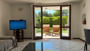 Sala de estar con televisor, sofá, mesa de centro y puerta corrediza de vidrio que da a un patio con sillas y vistas a zonas verdes.