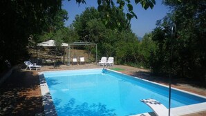 A rectangular swimming pool with a clear blue water surface, surrounded by a tiled deck. There are white lounge chairs and a covered seating area with a table and chairs in the background.