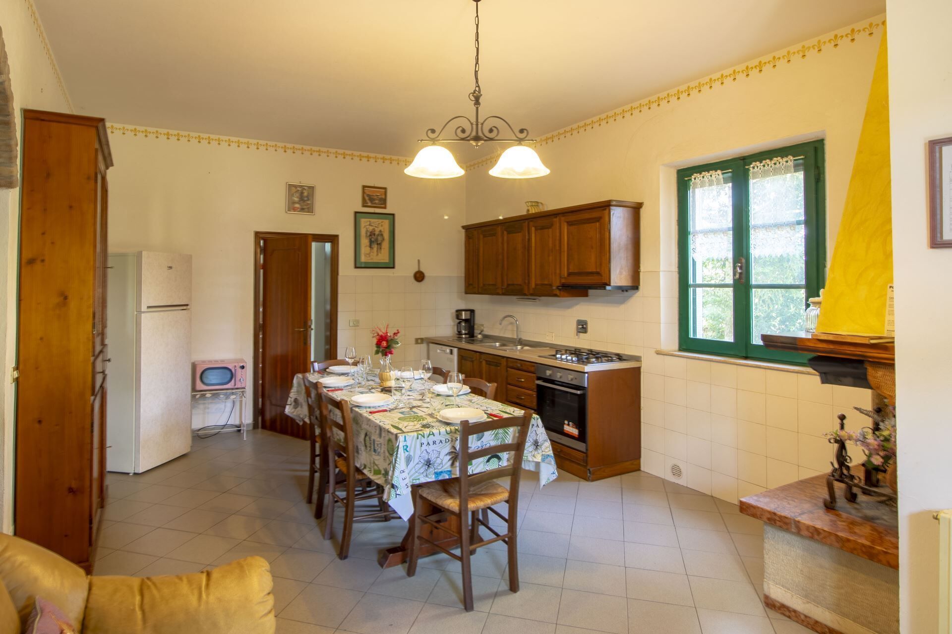 A kitchen with wooden cabinets, a dining table set for four, a refrigerator, and a window with a curtain.