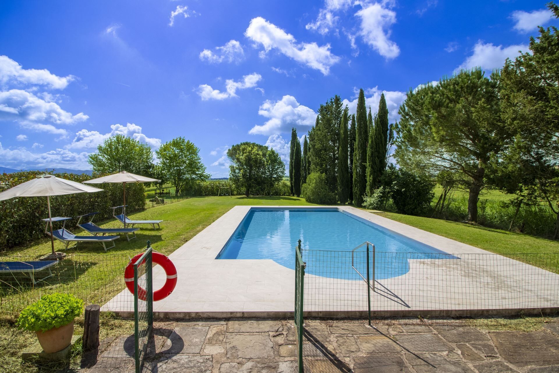 A rectangular swimming pool with a safety fence, surrounded by a grassy area with trees and a clear blue sky.