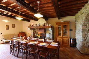 A dining area with a wooden table set for four, surrounded by chairs. There is a stone wall and a wooden cabinet with glass doors.