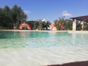 A clear swimming pool with a stone border, surrounded by lush greenery and a red building with a white dome. - Idyllic Trullo with salt water pool, Nestled Amongst  Ancient Olive Groves. (San Vito Dei Normanni)