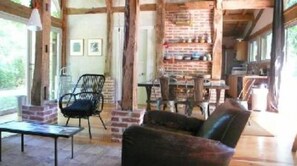 Een rustiek interieur met zichtbare houten balken, een metselstenen openhaard, een eethoek met tafel en stoelen, en een zithoek met een bank en fauteuil.