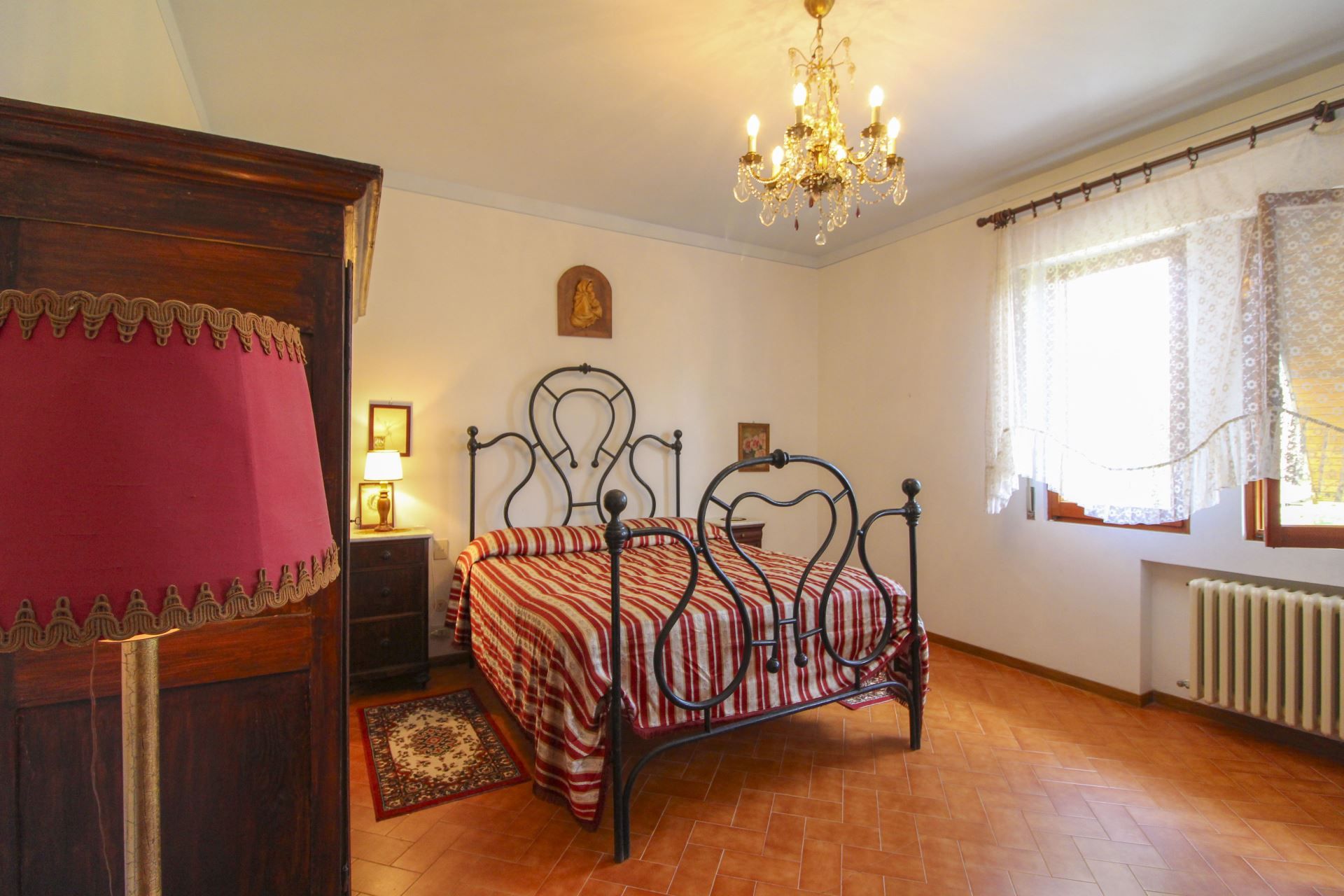 A bedroom with a bed, a nightstand, a dresser, a chandelier, and a window with curtains.
