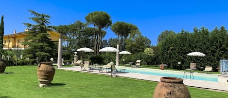 Zona de piscina con sillones de descanso y sombrillas, rodeada de vegetación y un edificio.