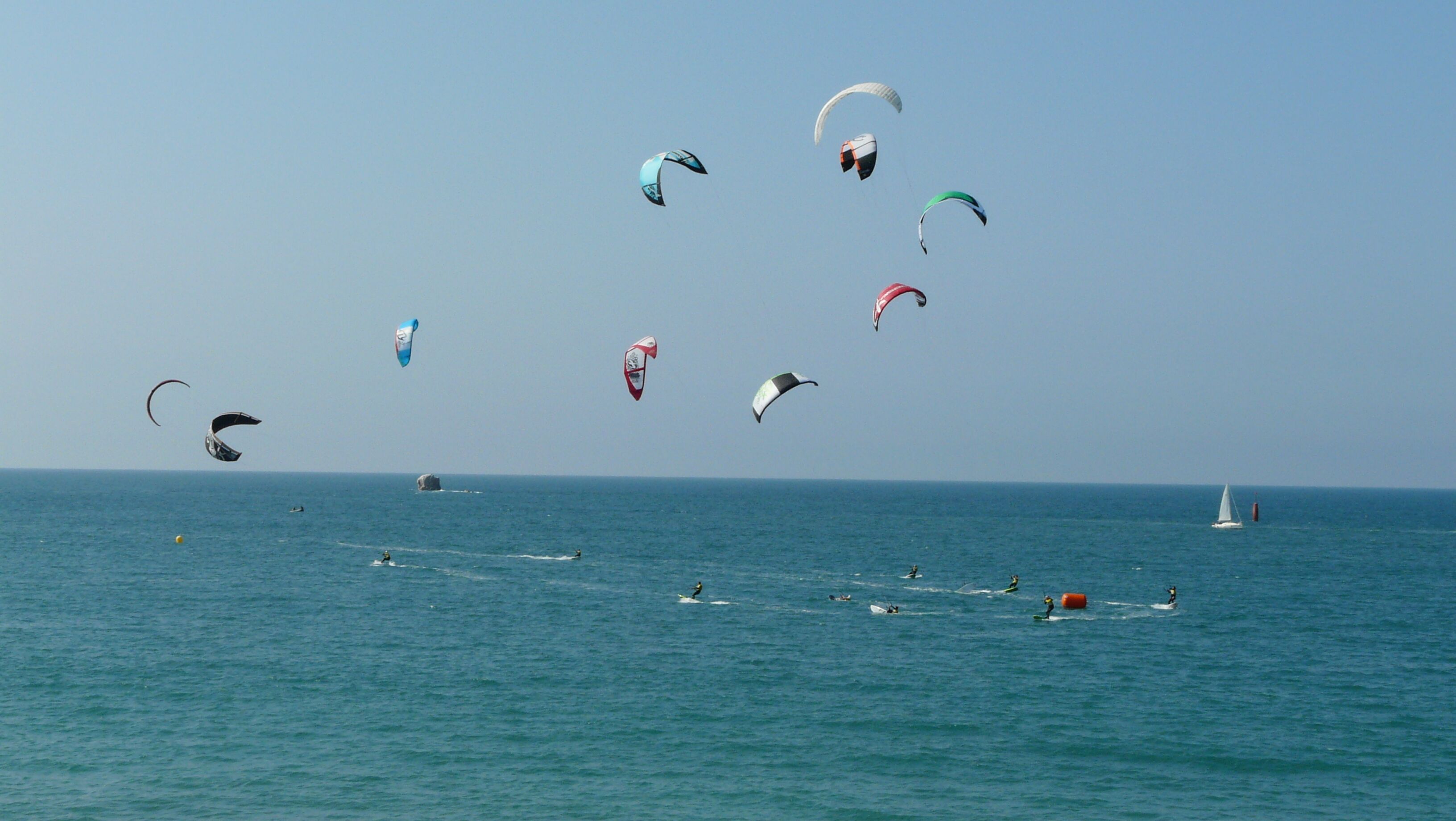 Une scène côtière avec plusieurs pratiquants de kitesurf dans l'eau et des cerfs-volants dans le ciel.
