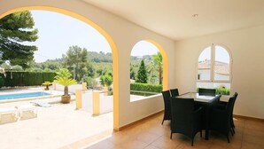 A spacious room with a tiled floor, a dining area with chairs, and a view of a swimming pool and greenery outside. - Caleta - Javea, Costa Blanca (Javea)