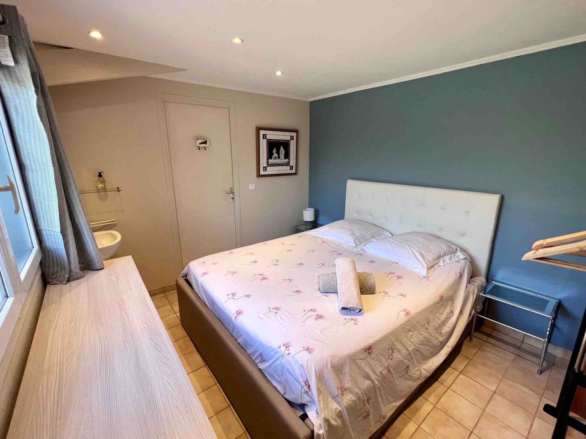 A bedroom with a bed, a bedside table, a window with curtains, a bathroom with a sink, and a framed picture on the wall.