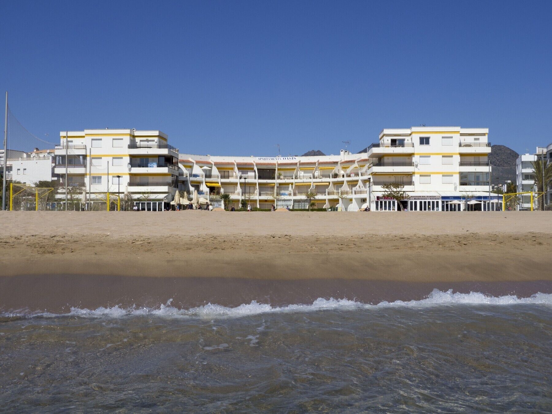 Un hotel frente a la playa con varios balcones, una playa arenosa en primer plano y cielos despejados.