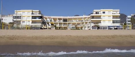 Un hotel frente a la playa con varios balcones, una playa arenosa en primer plano y cielos despejados.