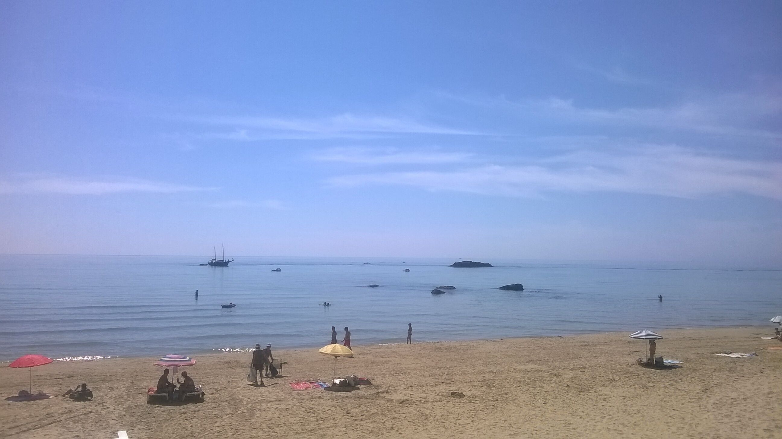 A beach with several boats in the water, people walking along the shore, and beach umbrellas.