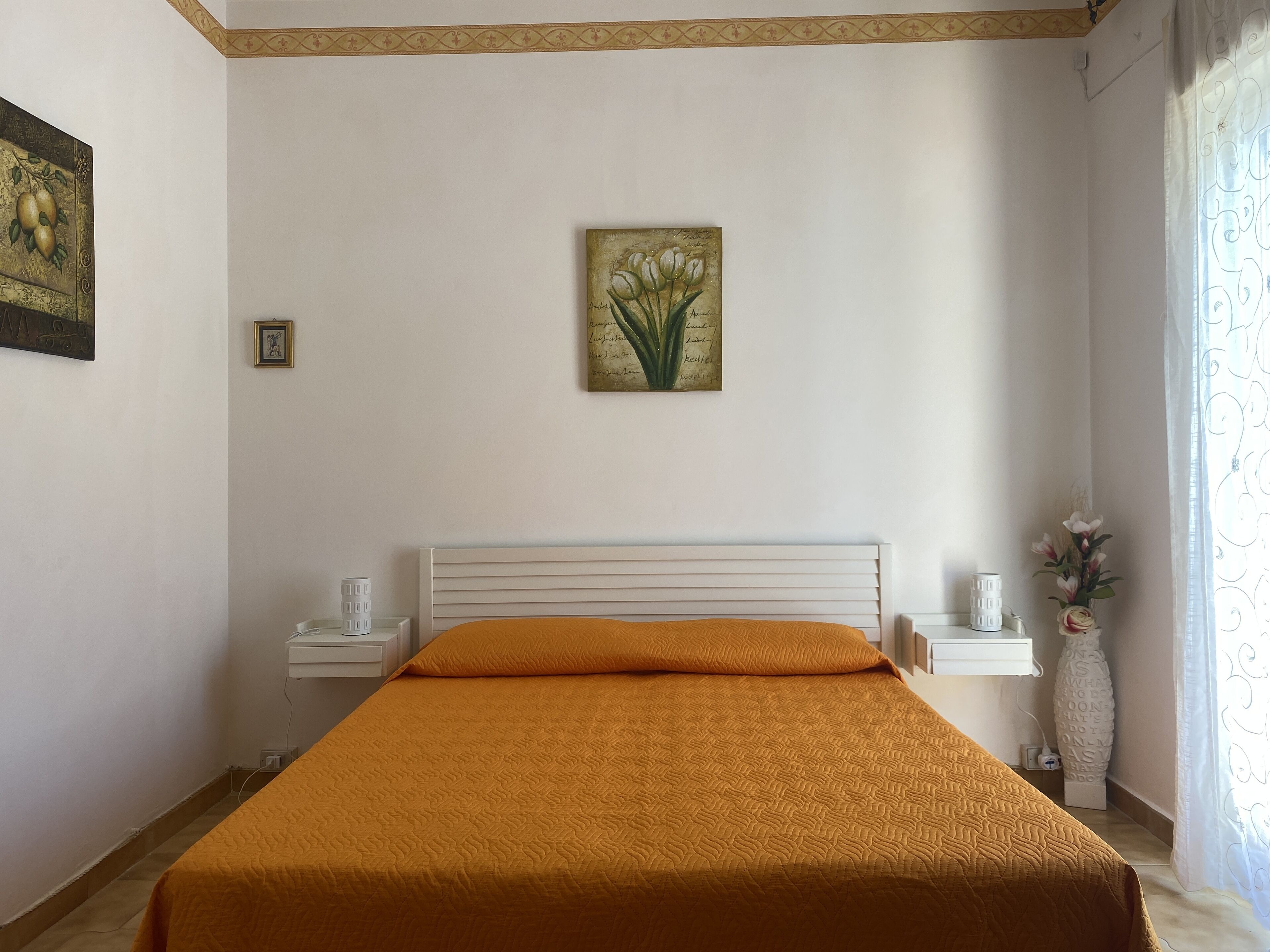 Una camera da letto con un letto, un comodino, un quadro appeso al muro e un vaso con fiori.