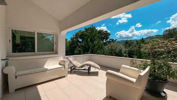 A balcony with a white sofa and two armchairs, a potted plant, and a view of trees and mountains.