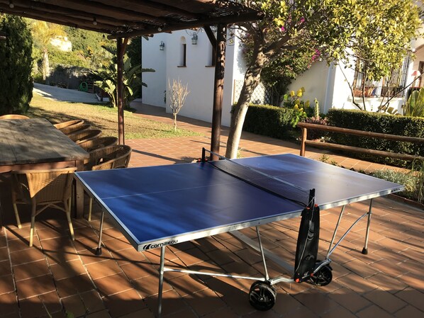 Una mesa de ping pong con ruedas, bajo una pérgola, en un patio con un edificio blanco al fondo.