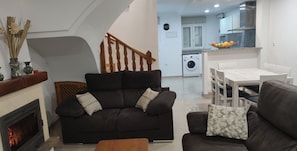 Una sala de estar moderna con un sofá negro, una mesa de centro de madera y una chimenea. Una escalera conduce a una zona de cocina con lavadora y una mesa de comedor con sillas.