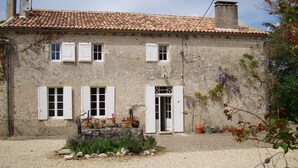 Un bâtiment en pierre de deux étages, avec un toit en tuiles, des volets blancs et un petit jardin agrémenté de plantes en pot.