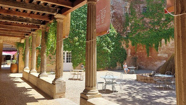 Un espace extérieur abrité, avec des colonnes en pierre, des tables et des chaises, et un bâtiment dont les murs sont recouverts de lierre, visible à l’arrière-plan.