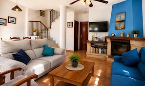 Un salon avec un canapé, une table basse et une cheminée. On y trouve un escalier avec une rampe et un téléviseur fixé au mur.