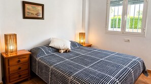 Une chambre à coucher comprenant un lit, deux tables de chevet, une commode, une lampe fixée au mur, une fenêtre avec des stores et un tableau encadré accroché au mur.