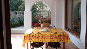 Eine Terrasse mit einem Tisch für zwei Personen, bedeckt mit einer gelb-rot karierten Tischdecke, zwei schwarzen Metallstühlen und einer weißen Vase mit gelben Blumen.