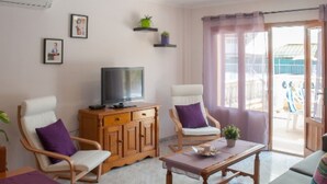 Un salón con televisor, dos sillones blancos con cojines morados, un aparador de madera y una mesa con una planta.