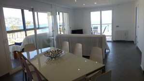 Un espace repas moderne avec une table et des chaises, de grandes fenêtres donnant sur la mer et un lit en arrière-plan.