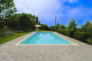 A rectangular swimming pool with a stone deck, surrounded by a hedge and a row of sun loungers.