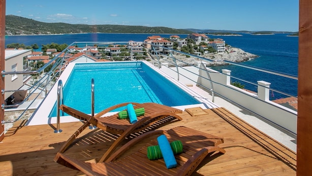Une terrasse en bois avec une piscine et des chaises longues, offrant une vue sur une ville côtière et la mer.