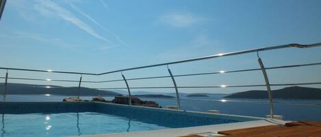 A pool with a wooden deck, surrounded by metal railings, overlooking a body of water with distant landforms.