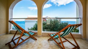 Deux chaises pliantes en bois, dotées de coussins à rayures vertes et blanches, sont placées sur un sol carrelé, face à un balcon d’où l’on peut admirer la mer et les bâtiments.