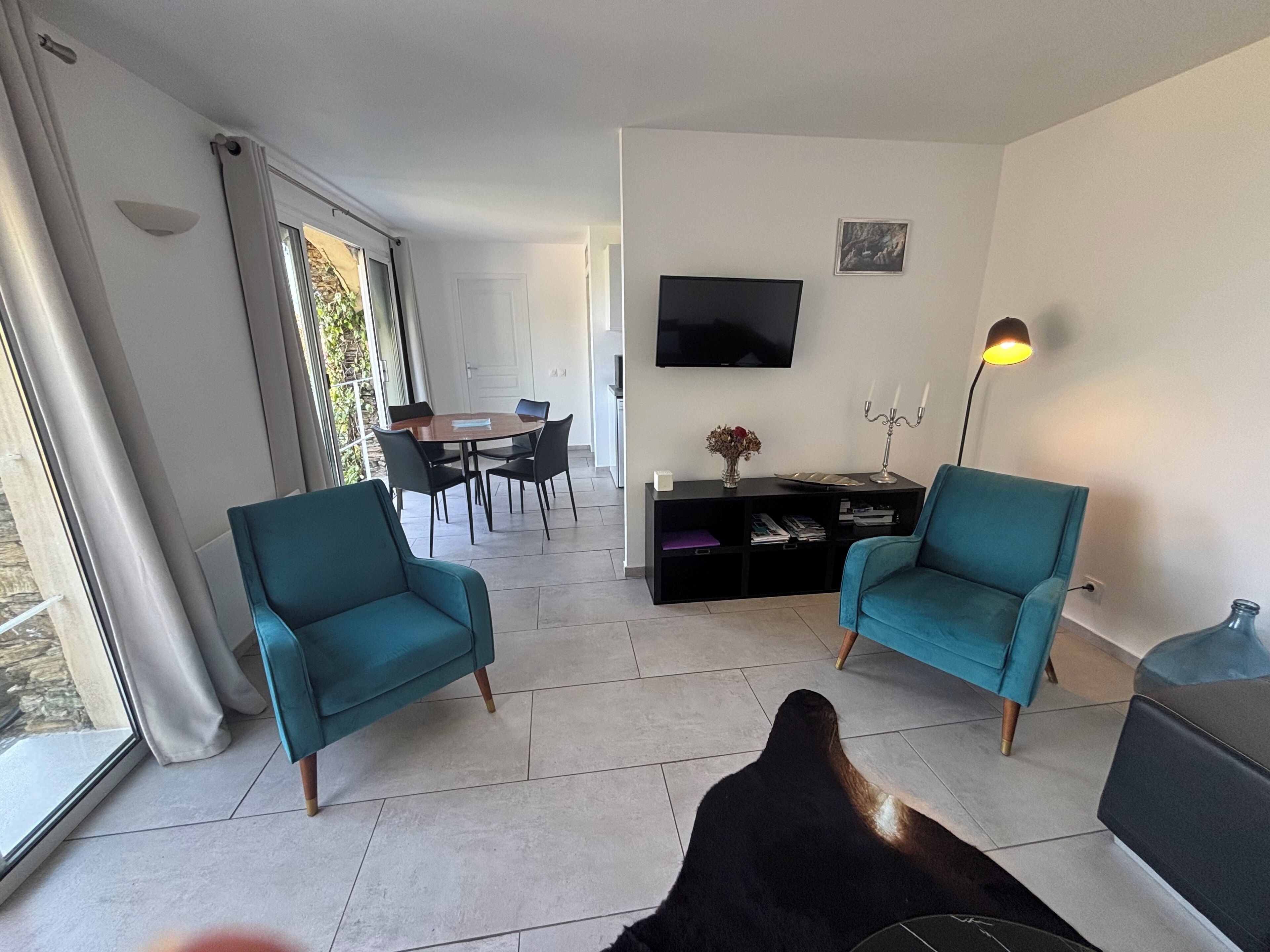 Un salon moderne avec des fauteuils turquoise, un téléviseur à écran plat, un coin repas avec une table et des chaises, et une grande fenêtre.