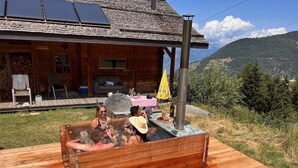 A wooden hot tub with people relaxing, a wooden cabin with a sloped roof, and a mountainous backdrop. - La Tzoumaz, 'BeauChalet'. Large 8-person chalet, Ski In, ski out (La Tzoumaz)
