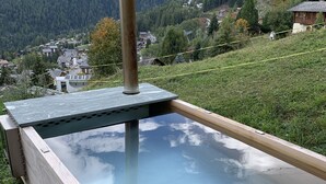 A wooden hot tub with a metal heater, surrounded by a deck, set against a mountainous backdrop. - La Tzoumaz, 'BeauChalet'. Large 8-person chalet, Ski In, ski out (La Tzoumaz)