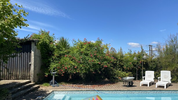 Une piscine avec un toboggan aux couleurs vives, des chaises longues blanches et un jardin agrémenté d’une clôture en bois et d’arbustes en fleurs.